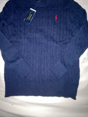 Mörkblå kabelstickad tröja Polo Ralph Lauren - Kabelstickad mörkblå tröja från Polo Ralph Lauren med rund halsringning och röd broderad logga på bröstet. Tröjan har ribbade muddar vid ärmslut och nederkant. Perfekt för dig som gillar klassisk och stilren design.