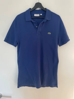 Lacoste pikétröja – blå – Slim Fit – M - Stilren pikétröja från Lacoste i blå färg. Klassisk modell med broderad krokodil på bröstet. Perfekt till både vardag och sommaroutfits.  ✔ Storlek: M (Slim Fit) ✔ Färg: Blå ✔ Material: 100% bomull ✔ Skick: Bra skick