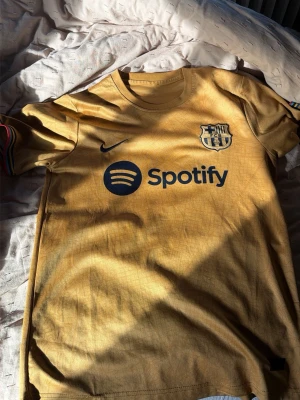 Gul FC Barcelona matchtröja Nike - Snygg gul FC Barcelona matchtröja från Nike med klubbmärke och Spotify-logga på bröstet. Tröjan har korta ärmar och rund hals, samt diskreta ränder i klubbens färger på ärmsluten. Perfekt för dig som älskar fotboll och vill representera Barca.