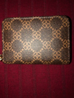 Brun mönstrad plånbok med dragkedja - Snygg brun plånbok med guldfärgad dragkedja och grafiskt mönster i beige. Plånboken har flera fack på insidan för kort och sedlar. Tillverkad i konstläder med textilfoder. Perfekt storlek för att ha med sig i fickan eller väskan.