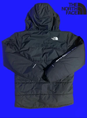 Svart pufferjacka från The North Face - Snygg svart pufferjacka från The North Face med huva och dragkedja framtill. Jackan har logga på bröstet och ärmen, samt diskreta reflexdetaljer. Tillverkad i slitstarkt syntetmaterial som håller värmen under kalla dagar.