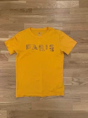 Gul Paris t-shirt från Jordan - Snygg gul t-shirt från Jordan med trycket 'PARIS' i svart på bröstet och små detaljer som Jordan-loggan och PSG-emblemet. Rund halsringning och korta ärmar. Perfekt för dig som gillar streetwear och sportiga vibes.