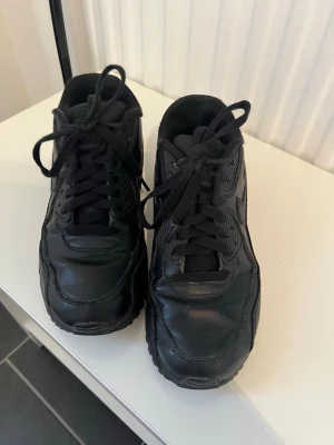 Nike Air Max sneakers - Säljer ett par klassiska Nike Air Max 90 sneakers i helsvart läder med matchande svarta skosnören och sula. Ikonisk Nike Swoosh på sidan och Air Max-detalj vid hälen. Perfekta för dig som gillar en clean och tidlös look.