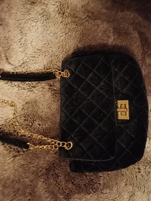 Svart axelväska med guldkedja - Snygg svart axelväska med guldfärgad kedja. Väskan har en klassisk, rektangulär form. Får plats med de viktigaste 