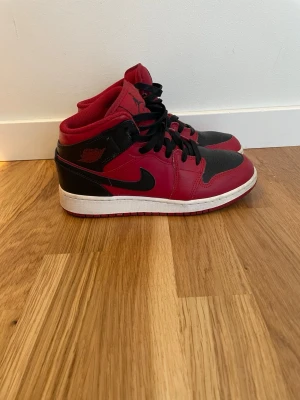 Air Jordan 1 Mid röd/svart - Snygga Air Jordan 1 Mid sneakers i rött och svart med klassisk Nike Swoosh på sidan. Skorna har svart snörning, vit mellansula och röd yttersula. Tillverkade i läder och textil, med ikoniska Jordan-loggor på plös och vid ankeln. Perfekta för dig som gillar streetwear.