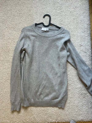 Ljusgrå stickad tröja från Vila - En enkel och stilren ljusgrå stickad tröja från Vila. Tröjan har rund halsringning, långa ärmar och ribbade muddar vid ärmslut och nederkant. Perfekt basplagg att matcha med jeans eller kjol. Materialet är mjukt och bekvämt.