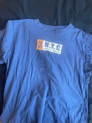 Blå NYC t-shirt med tryck - Blå t-shirt med tryck framtill där det står 'N.Y.C' och '1999' i vitt och orange. T-shirten har rund hals och korta ärmar, perfekt för en avslappnad streetwear-look. Tillverkad i mjuk bomull för skön känsla hela dagen. (klicka ej köp nu ) kontakta gärna och Swish går ❤️