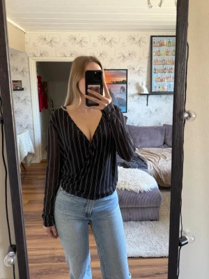 Svart omlottblus med vita ränder - Snygg svart omlottblus med tunna vita vertikala ränder och djup v-ringning. Blusen har långa ärmar och ett avslappnat fall. Perfekt att styla med jeans eller kjol för en trendig look. Den är från NAKD storlek xs men mer som s/m, använd en tån