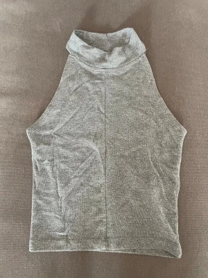 Glittrig grå polotopp - Glittrig grå polotopp med hög krage och ärmlös design. Toppen har en mjuk och stretchig känsla, perfekt för att lyfta vilken outfit som helst. Materialet skimrar snyggt i ljuset 