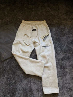 Essentials Fear of God mjukisbyxor grå - Snygga ljusgrå mjukisbyxor från Essentials Fear of God med dragsko i midjan och resår vid benslut. Byxorna har en stilren logga på benet och är tillverkade i mjukt material som passar perfekt för chill dagar. Kommer med originalpåse och tag kvar.