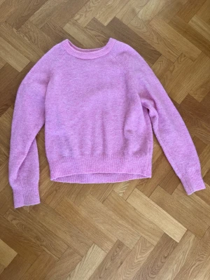 Ljusrosa stickad tröja från C.W. - Mysig ljusrosa stickad tröja från C.W. med rund halsringning och ribbade muddar vid ärmslut och nederkant. Tröjan har en enkel, klassisk design och är gjord i ett mjukt material som känns skönt mot huden. Perfekt för lager på lager under kyliga dagar.