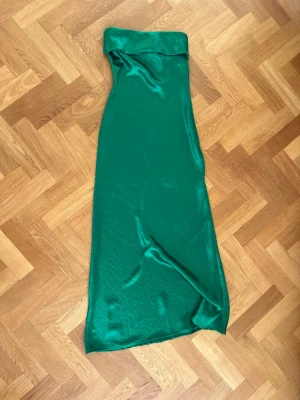 Grön axelbandslös långklänning - Säljer en smaragdgrön axelbandslös långklänning från In the style i glansigt satinliknande material. Klänningen har en elegant, figurnära passform och släpper mjukt längs kroppen. Perfekt för dig som vill sticka ut med en färgstark look.