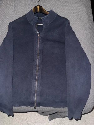 Mörkblå ziptröja Massimo Dutti - Snygg mörkblå ziptröja från Massimo Dutti med hög krage och hel dragkedja framtill. Tröjan har ribbade muddar vid ärmslut och nederkant, vilket ger en clean och stilren look. Perfekt att slänga på sig över en t-shirt för en avslappnad men ändå uppklädd stil.