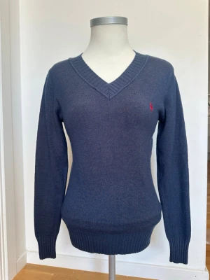 Mörkblå stickad tröja från Ralph Lauren - Mörkblå stickad tröja från Ralph Lauren med V-ringning och ribbade muddar. Tröjan har en liten röd broderad logga på bröstet och är tillverkad i ett mjukt, tunt material. Perfekt för lager på lager och enkel att matcha.