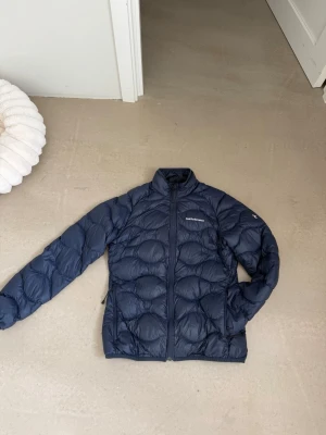 Mörkblå dunjacka från Peak Performance - Snygg mörkblå dunjacka från Peak Performance med hög krage och dragkedja framtill. Jackan har ett quiltat mönster och två sidofickor med dragkedja. Perfekt för kalla vinterdagar och riktigt lätt att bära.