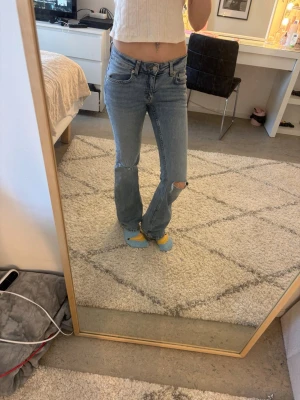 Ljusblå bootcut jeans med slitning - Säljer ett par ljusblå bootcut jeans med slitning på ena knät. Jeansen har klassisk femficksmodell, normal midja och är tillverkade i denim. Perfekta för en avslappnad och trendig look.