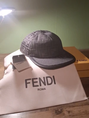 Svart keps med FF-mönster från Fendi - Snygg svart keps från Fendi med det klassiska FF-monogrammet över hela kepsen. Skärmen är i svart läderlook och kepsen har en modern, rundad form. Perfekt för dig som vill ha en exklusiv accessoar med streetkänsla. HELT OANVÄND DU FÅR ALLT I BILDEN!!!