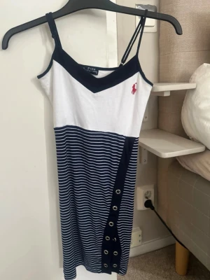 Randig klänning från Polo Ralph Lauren - Snygg kort klänning med smala axelband från Polo Ralph Lauren. Överdelen är vit med marinblå detaljer och logga, nederdelen är randig i vitt och marinblått med dekorativa knappar längs ena sidan. Perfekt för sommarens alla tillfällen.