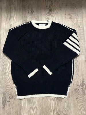 Thom Browne stickad tröja  - Snygga mörkblåa stickade tröja från Thom Browne med cremfärgade detaljer och fyra ränder på ena ärmen. Tröjan har ribbade muddar, knappar i sidorna och röd-vit-blåa band på insidan. Perfekt för dig som gillar klassisk men unik stil. Står storlek S men passar som M