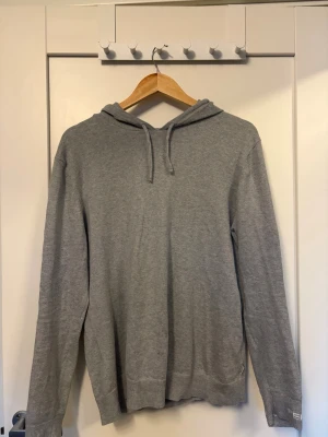 Grå hoodie med dragsko - En stilren grå hoodie med huva och dragsko. Tillverkad i mjukt, tunt material som känns skönt mot huden. Klassisk passform med ribbade muddar vid ärmslut och nederkant. Perfekt för en avslappnad look.