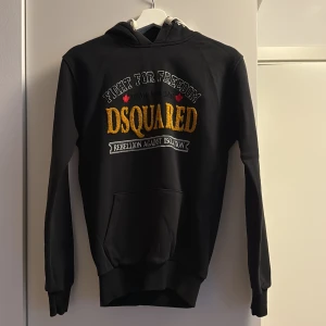 Svart hoodie från Dsquared2 - Svart hoodie från Dsquared2 med broderad text i gult, vitt och rött på bröstet. Snygg huva med vita snören och stor magficka framtill. Tillverkad i mjukt material, perfekt för en avslappnad streetwear-look.