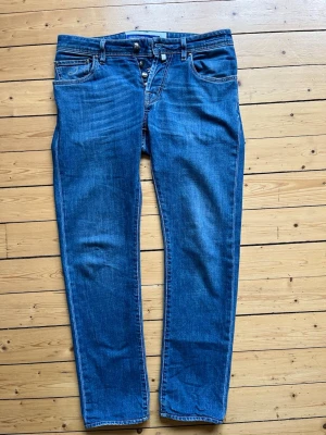 Jacob Cohen blå jeans  - Jacob Cohen jeans i modellen Nick. Jeansen har en knappgylf och detaljerade sömmar. Bakfickorna har Jacob Cohens signaturdesign och en grön läderlapp med logotypen. Storlek 30 Längd 96cm Bredd 44cm