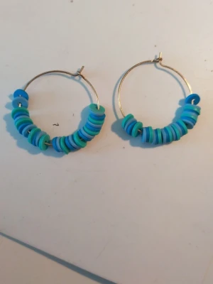 Hoops med blå och turkosa pärlor - Säljer ett par coola örhängen i form av hoops med blå och turkosa platta pärlor. Örhängena har en enkel metallring som bas och pärlorna är staplade tätt intill varandra för en färgglad look. Perfekta för att addera lite färg till din stil!