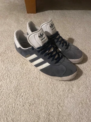 Adidas Gazelle navy/vita sneakers - Snygga Adidas Gazelle sneakers i navy mocka med vita detaljer och klassiska tre ränder på sidan. Skorna har vit sula, vit hälkappa och mörkblå skosnören. Perfekta för en stilren och sportig look.