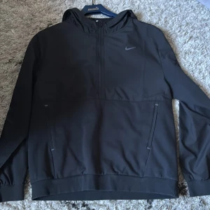 Svart hoodie från Nike med dragkedja - Svart hoodie från Nike med halv dragkedja framtill och diskret logga på bröstet. Modellen har huva, långa ärmar och två snedställda fickor med dragkedja. Perfekt för en sportig och avslappnad stil.