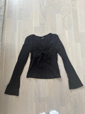 Svart långärmad topp med knyt - Snygg svart långärmad topp med v-ringning och knyt framtill. Ärmarna är långa och lätt utsvängda, vilket ger en trendig look. Perfekt att styla med jeans eller kjol för en cool outfit. Ny utan prislapp 