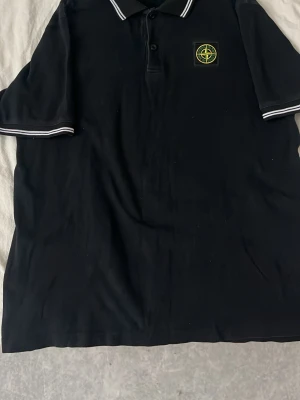 Svart pikétröja från Stone Island - Klassisk svart pikétröja från Stone Island med vit piping på krage och ärmslut. Tröjan har knappslå framtill och den ikoniska Stone Island-loggan på bröstet. Tillverkad i bomull för en skön och avslappnad känsla.