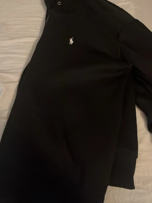 Svart zip-jacka från Polo Ralph Lauren - Snygg svart zip-jacka från Polo Ralph Lauren med klassisk vit broderad logga på bröstet. Jackan har hög krage, muddar vid ärmslut och nederkant samt fickor på sidorna. Perfekt för en stilren och avslappnad look. Äkta. Knappt använt. Inga problem med den.
