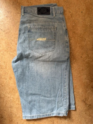Ljusblå jeansshorts från Joker Brand s38 - Säljer ett par ljusblå jeansshorts från Joker Brand med klassisk femficksdesign och logga på bakfickan. Shortsen har normal passform och är tillverkade i bomull. Perfekta för en avslappnad stil under varmare dagar.