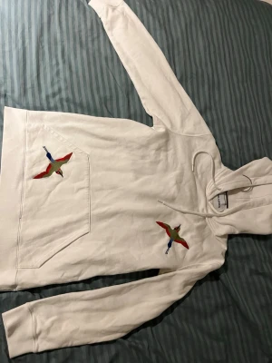 Vit hoodie med broderade fåglar - Säljer en vit hoodie med huva och dragsko. På bröstet och fickan finns färgglada broderade fåglar i rött, grönt och blått. Hoodien har en stor magficka och är gjord i mjukt bomullstyg. Perfekt för dig som gillar unika detaljer och streetwear-vibe.