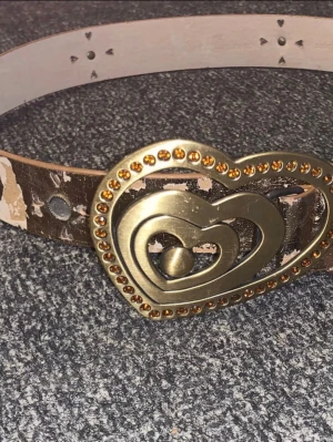 Brunt bälte med hjärtspänne - Snyggt brunt bälte med ett stort hjärtformat spänne i guld och nitar runt kanten. Bältet har en sliten look och passar perfekt för att ge outfiten en unik touch. Materialet är skinn och spännet är i metall.