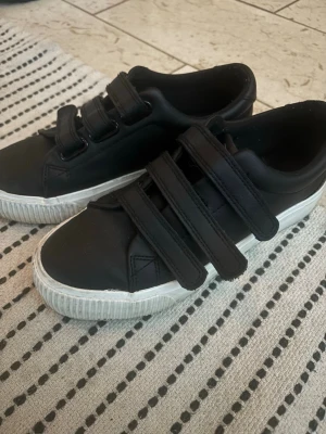 Svarta sneakers från Even&Odd - Snygga svarta sneakers från Even&Odd med tre breda kardborreband och vit sula. Skorna har en klassisk låg modell och är tillverkade i ett slätt syntetmaterial. Perfekta för en clean och enkel stil.