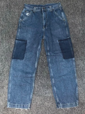 Baggy jeans med patchade knän - Säljer ett par baggy blå jeans med stora mörkblå patchar på knäna och raka ben. Jeansen har klassiska fickor fram och bak, samt en vit logga på ena bakfickan. Perfekta för en avslappnad streetstil.