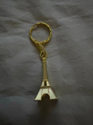 Guldfärgad Eiffeltorn nyckelring, köpt i Paris - Köpt i Paris. Snygg nyckelring i metall med en detaljrik Eiffeltorn-berlock i guldfärg. Perfekt accessoar för dig som gillar Paris eller vill ha något unikt till dina nycklar. Liten och smidig att ha i fickan eller väskan.