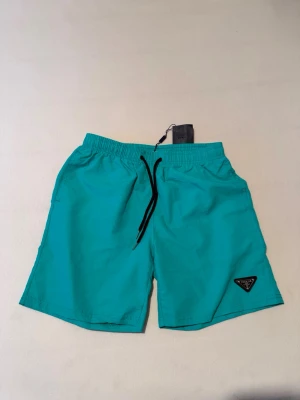 Turkosa badbyxor med svart snöre - Snygga turkosa badbyxor med svart dragsko i midjan och elastisk resår. Klassisk modell med fickor och en liten svart logotyp på vänster ben. Perfekta för stranden eller poolen, tillverkade i snabbtorkande material.