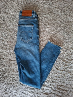 Blå Tiger Of Sweden skinny jeans med rå kant - Säljer ett par blå skinny jeans med hög midja och rå, fransig kant vid bensluten. Jeansen har klassiska fem fickor, dragkedja och knapp framtill samt en snygg tvätt med lätt slitning. Perfekta för en trendig och avslappnad look.