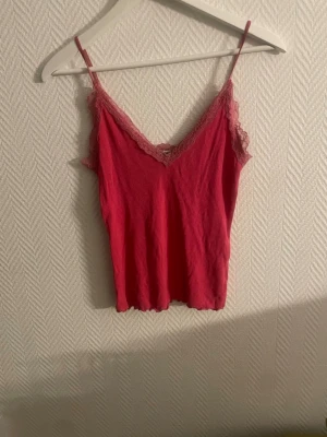 Rosa ribbad linnetopp med spets - Säljer en söt rosa linnetopp med tunna axelband och v-ringning. Toppen har ribbad struktur och spetsdetaljer längs urringningen och ärmarna för en extra feminin touch. Perfekt att styla med jeans eller kjol för en trendig look.