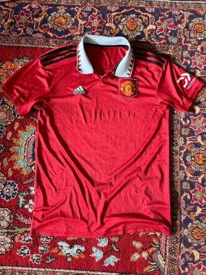 Manchester United röd matchtröja 2022/2023 - Säljer en röd Manchester United matchtröja från Adidas med klubbmärke på bröstet och vita detaljer på kragen. Tröjan har svarta ränder på axlarna och klassisk pikékrage. Perfekt för dig som älskar fotboll och vill visa ditt lagstöd.Tröjan är äkta och gå att verifiera det via numret i nacken, lite sliten på trycket på armen annars fint skick 