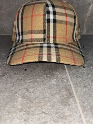 Beige rutig keps med klassiskt mönster - Säljer en beige keps med ikoniskt rutigt mönster i svart, vitt och rött. Kepsen har böjd skärm och justerbar baksida. Perfekt accessoar för dig som vill ha en stilren och tidlös look.