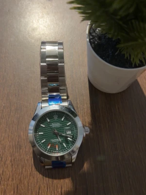 Klocka  - Stilren klocka med grön urtavla och silverfärgat armband i metall. Klockan har tydliga index, datumvisning och klassisk rund form. Perfekt accessoar för dig som vill sticka ut med en lyxig och tidlös look.