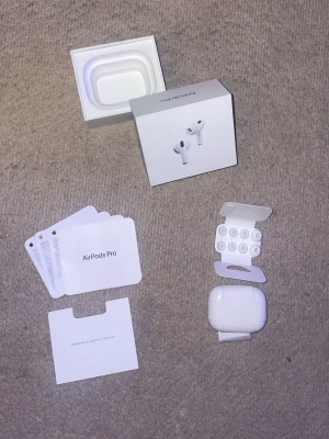Apple AirPods Pro (3:a gen) med tillbehör - Säljer ett par vita trådlösa in-ear hörlurar med laddningsetui. Hörlurarna har silikonproppar och en stilren, kompakt design. Perfekta för musik, poddar och samtal på språng. Laddningsetuiet har en LED-indikator på framsidan.