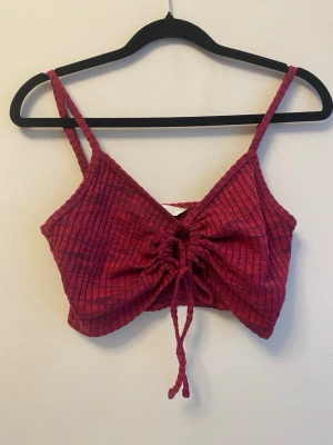 Vinröd ribbad croppad linnetopp - Trendig vinröd croppad linnetopp med ribbad struktur och justerbara axelband. Toppen har snygg snörning framtill som ger en cool detalj och markerar midjan. Perfekt för dig som gillar en stilren men ändå edgy look.