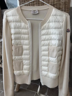 Beige quiltad cardigan från Moncler - Vit cardiganjacka från Moncler med quiltad framsida och stickade ärmar. Jackan har rund halsringning, dragkedja framtill och fyra små fickor på bröstet. Moncler-logga på ärmen. Nypris 9899kr. 
