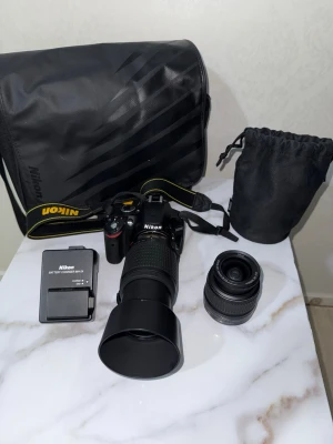 Nikon systemkamera med två objektiv - Säljer en svart Nikon systemkamera med två objektiv, kameraväska, batteriladdare och objektivpåse. Kameran har tydliga reglage och rem med Nikon-logga. Perfekt för n dig som vill ta steget in i systemkameravärlden och fota med olika brännvidder.Monterat teleobjektiv: Nikon AF-S DX Nikkor 55-200mm f/4-5.6G ED VR II.Motljusskydd: Ett runt, svart plastskydd sitter monterat längst fram på teleobjektivet.Löst standardobjektiv: Nikon AF-S DX Nikkor 18-55mm f/3.5-5.6G VR.Användning: 18-55mm är 