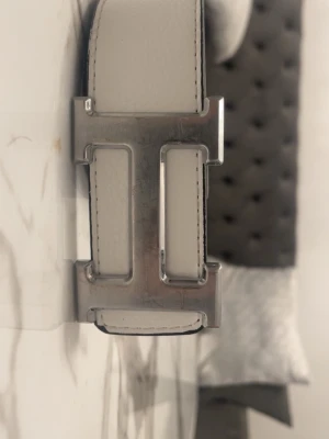 Vitt skärp med silverfärgat spänne - Snyggt vitt skärp i skinn med ett stort, stilrent silverfärgat spänne. Perfekt accessoar för att lyfta din outfit och ge en clean look. Skärpet har flera hål för justerbar passform och klassisk design. 110cm