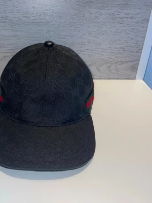 Svart keps med logga - Snygg svart keps med diskret monogram över hela kepsen och klassiska ränder på sidan. Kepsen har böjd skärm och knapp på toppen. Perfekt accessoar för en trendig streetwear-look.
